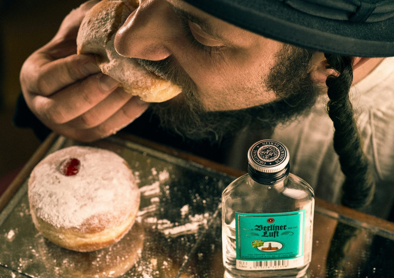 A person with a braided sidelock eats a powdered jelly donut beside another donut and a bottle labeled "Berliner Luft" on a reflective surface.