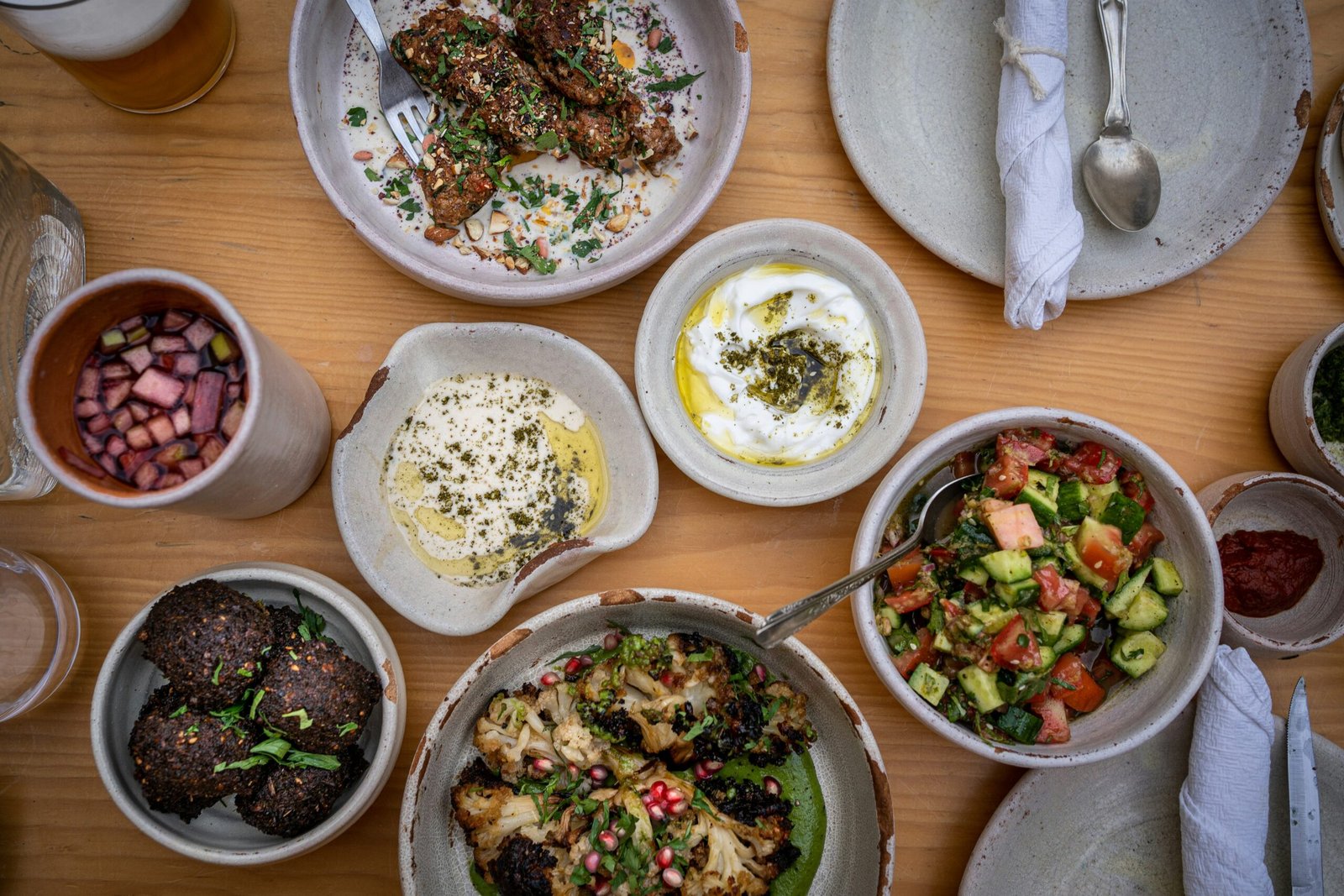 A variety of Middle Eastern dishes, including falafel, grilled vegetables, dips, salad, and a drink, are arranged on a wooden table with plates and utensils.